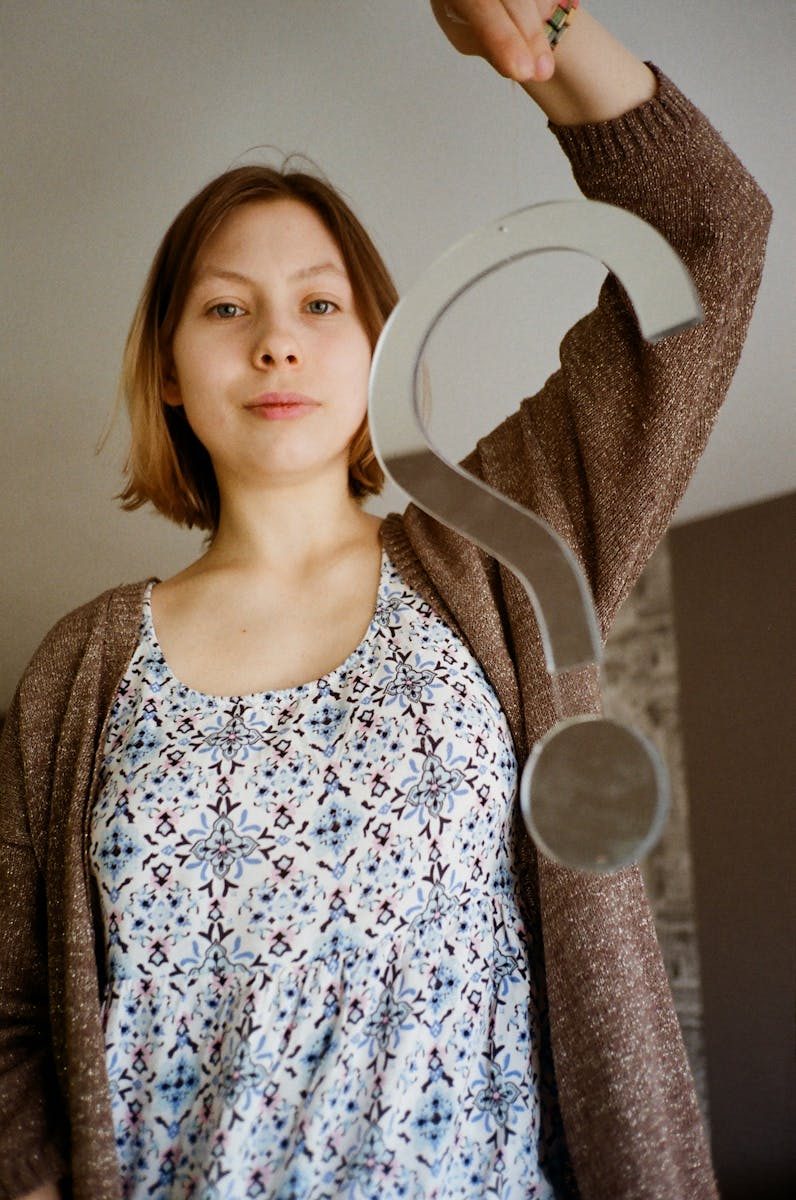 Caucasian woman holding a large question mark indoors, expressing curiosity.