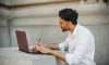 A man writing on a laptop outdoors, seated on a stone surface and focused on work.