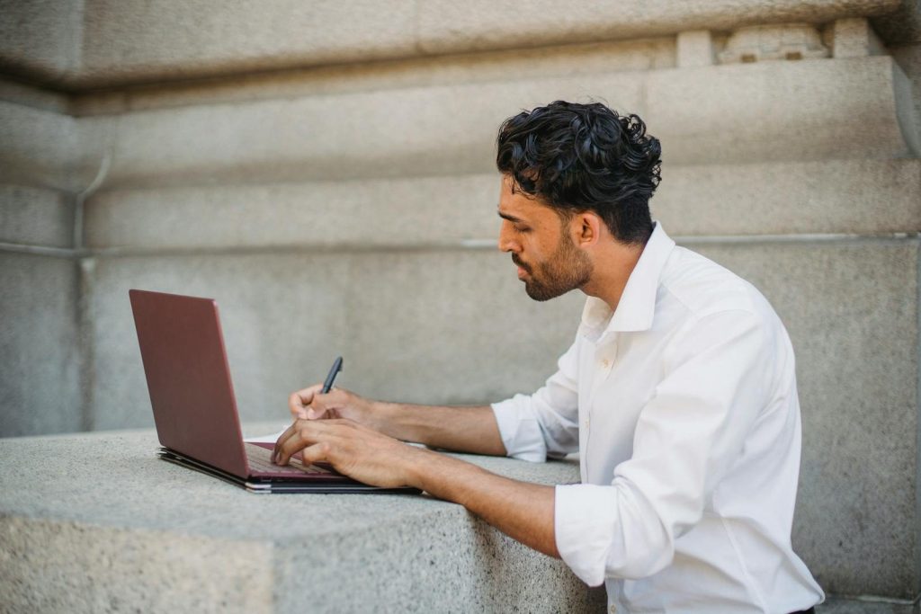 A man writing on a laptop outdoors, seated on a stone surface and focused on work.