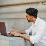 A man writing on a laptop outdoors, seated on a stone surface and focused on work.