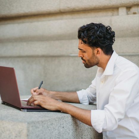 A man writing on a laptop outdoors, seated on a stone surface and focused on work.
