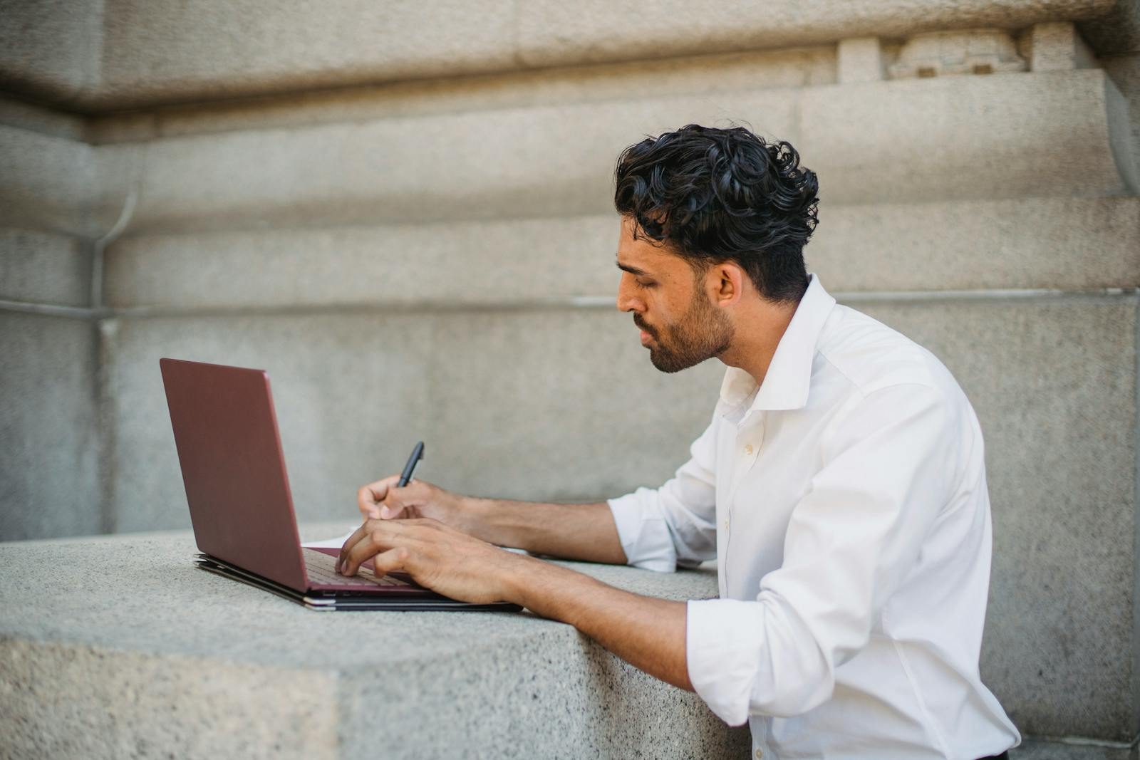 A man writing on a laptop outdoors, seated on a stone surface and focused on work.