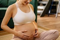 A smiling pregnant woman sits indoors, embracing her belly with love.