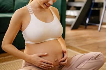 A smiling pregnant woman sits indoors, embracing her belly with love.