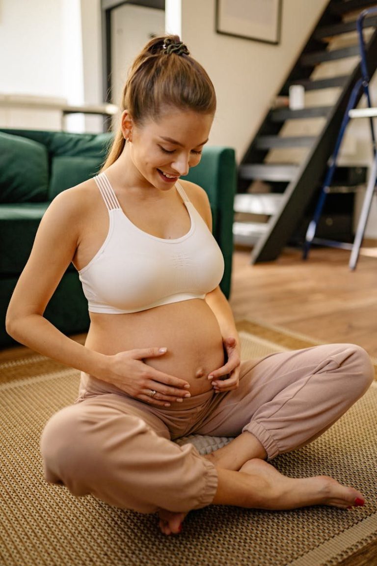 A smiling pregnant woman sits indoors, embracing her belly with love.
