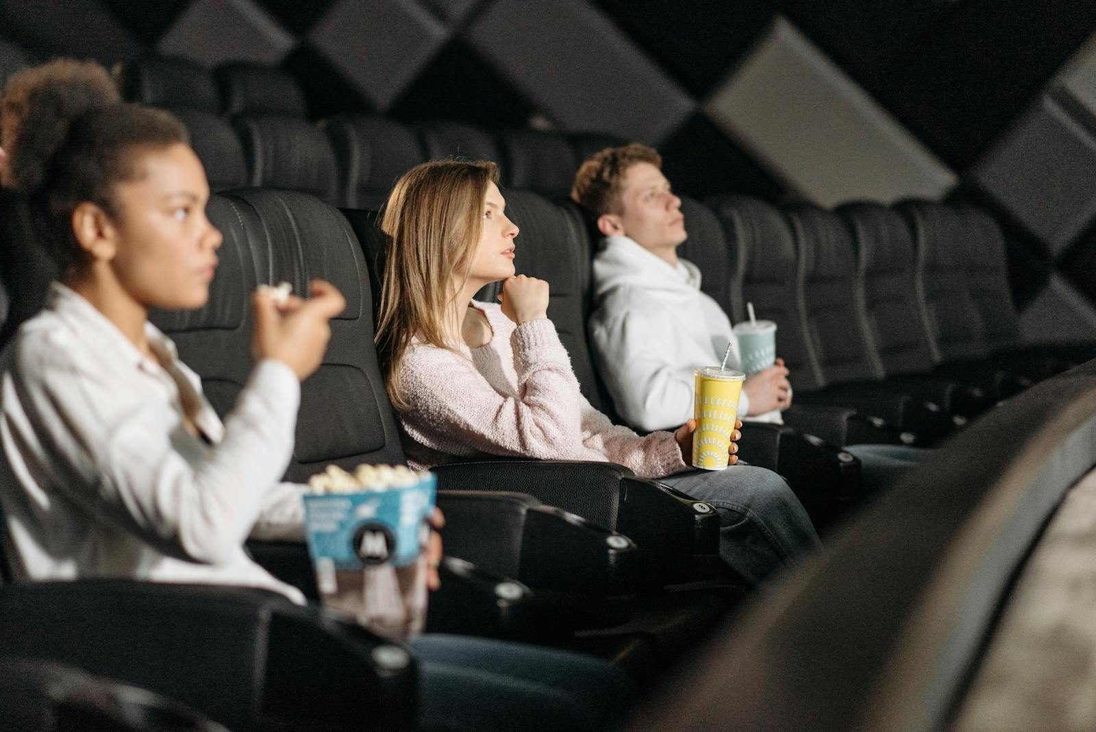 Three friends watching a movie while enjoying popcorn and drinks at a theater.