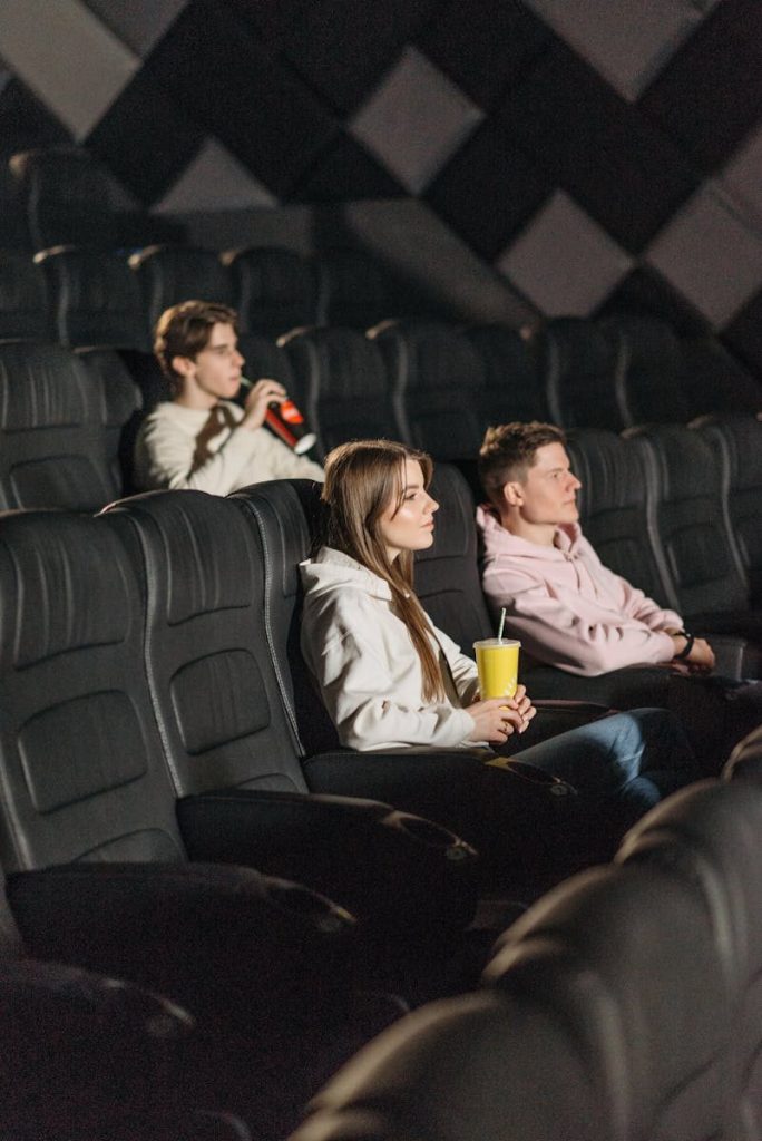 Three young adults seated in a movie theater enjoying a film. Holding snacks and drinks.