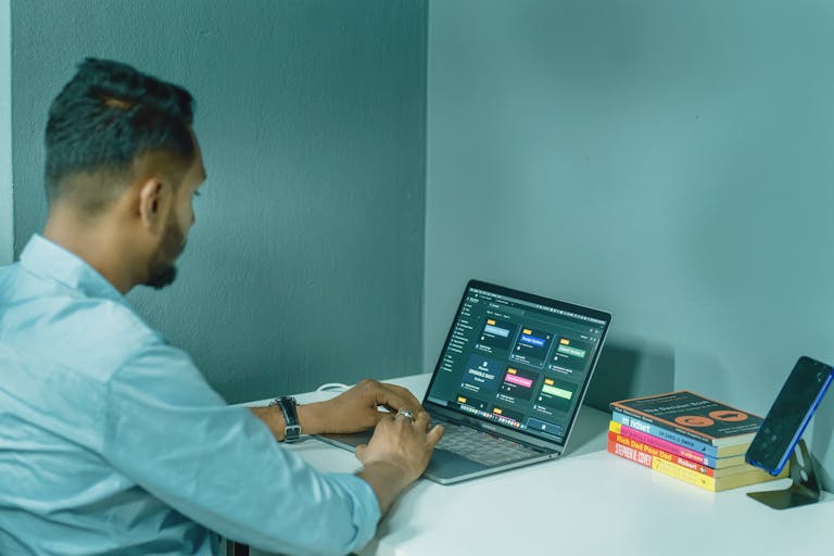 Focused man working on laptop at home office desk, surrounded by books and smartphone for a productive work environment.