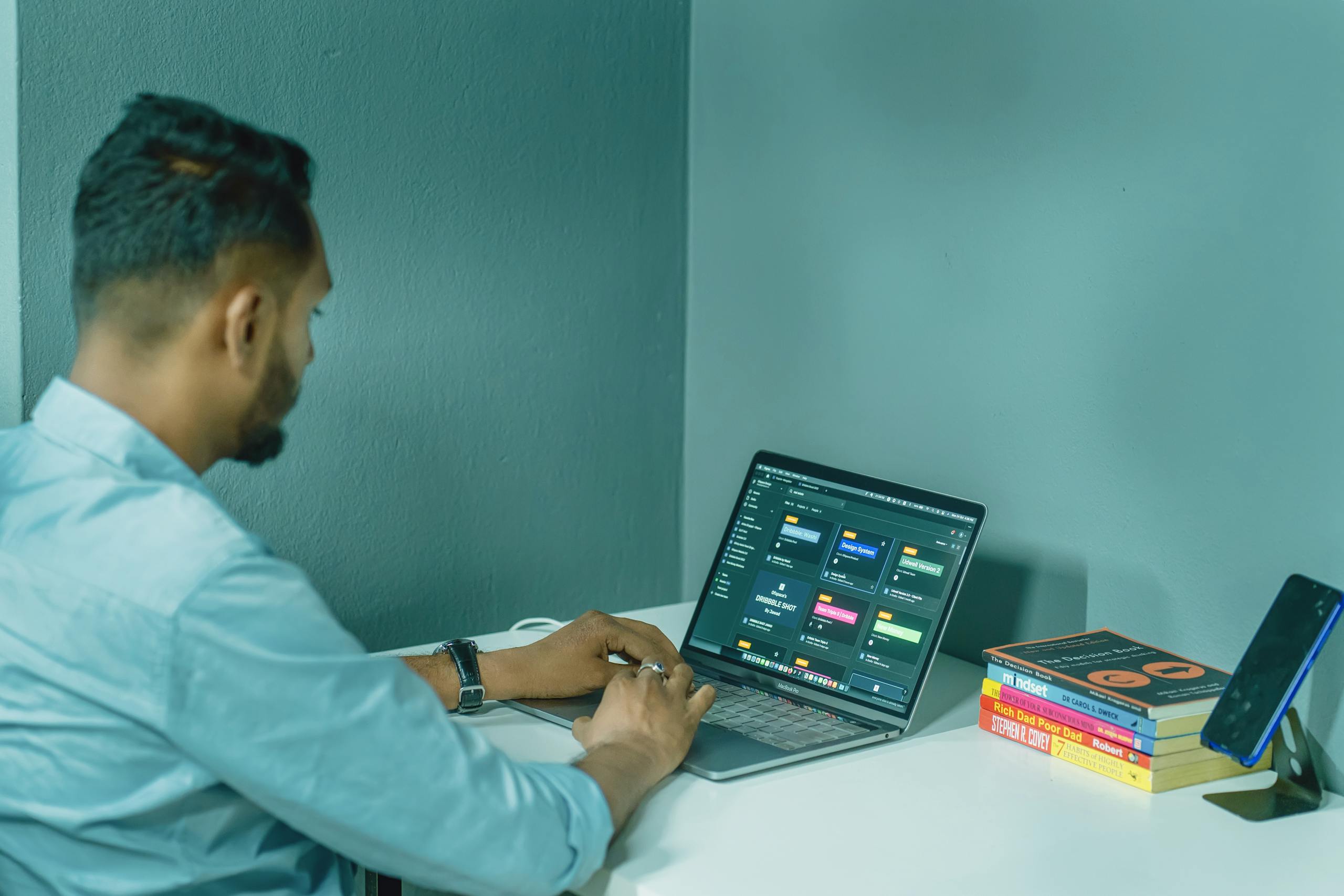 Focused man working on laptop at home office desk, surrounded by books and smartphone for a productive work environment.