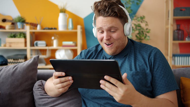 Smiling man with headphones using a tablet indoors, enjoying media content.