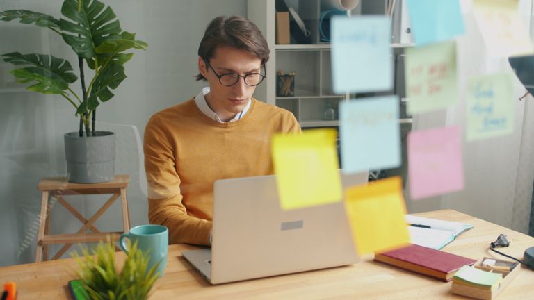 A young man focused on his laptop in a home office with sticky notes and plants.
