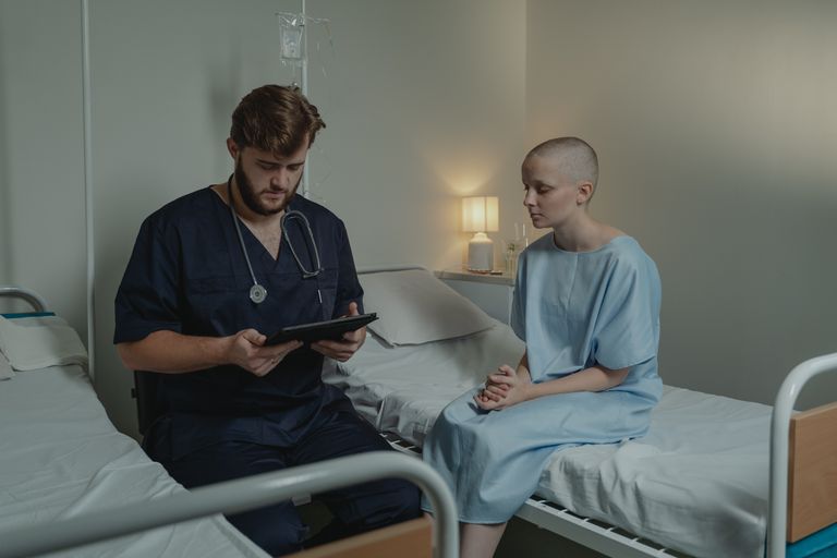 A doctor consulting a bald patient using a digital tablet in a hospital room.